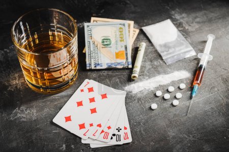 Boire de l'alcool dans un verre, jouer aux cartes pour le jeu de poker, seringue avec une dose de drogues, pilules blanches, narcotiques en poudre et la monnaie du dollar américain sur fond sombre. Concept de dépendance, de jeu et d'abus.