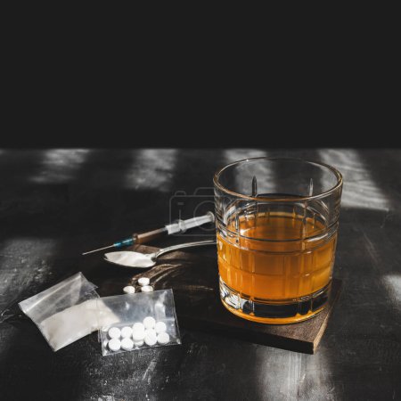 Foto de Beber alcohol en un vaso, jeringa con una dosis de drogas, pastillas blancas y polvo de narcóticos en una bolsa transparente sobre fondo oscuro. Concepto de adicción, abuso y malos hábitos. - Imagen libre de derechos