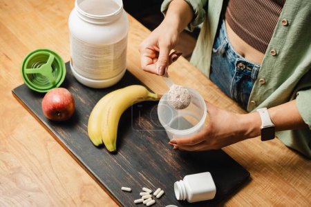 Photo for Woman in jeans and shirt with a measuring spoon in her hand puts portion of whey protein powder into a shaker on wooden table with white capsules, bananas and apple. Process of making protein drink. - Royalty Free Image
