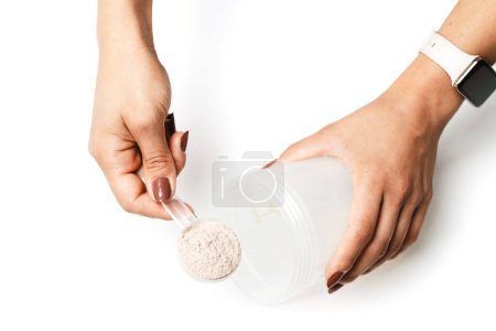 Photo for Woman's hands with measuring spoon puts portion of whey protein powder into a shaker, making protein drink cocktail, isolated on white background, top view. - Royalty Free Image