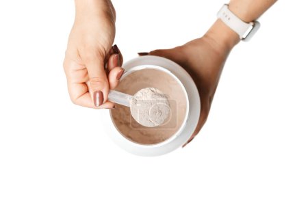 Photo for Woman's hand holding measuring spoon with portion of whey protein powder over a jar, making protein drink cocktail, isolated on white background, top view. - Royalty Free Image