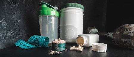 Chocolate whey protein powder in measuring spoon, white capsules of amino acids and vitamins, measuring tape, old rusty dumbbell, plastic shaker on dark background. bodybuilding food supplements.