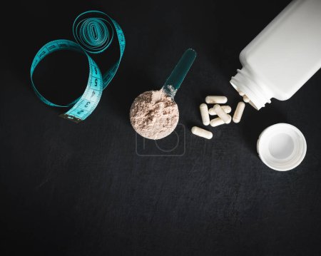 Photo for Chocolate whey protein powder in measuring spoon, white capsules of amino acids, vitamins and creatine, measuring tape, on dark background. bodybuilding food supplements, top view. - Royalty Free Image