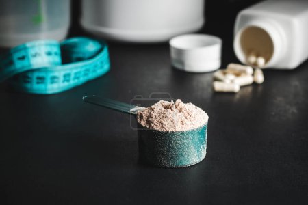 Photo for Chocolate whey protein powder in measuring spoon, white capsules of amino acids, vitamins and creatine, measuring tape, plastic shaker on dark background. bodybuilding food supplements. - Royalty Free Image