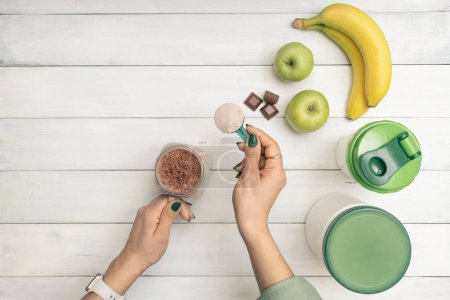 Photo for Young woman holding measuring spoon with protein powder, glass jar of protein drink cocktail, milkshake or smoothie above white wooden table with chocolate pieces, bananas and apples, top view. - Royalty Free Image