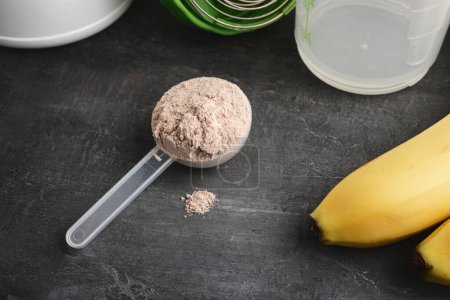 Proteína de suero en polvo con sabor a chocolate en una cuchara de plástico sobre un fondo oscuro con plátanos y una coctelera.