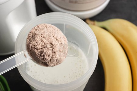 Cuchara de medición de plástico pone proteína de suero en polvo en una coctelera con leche sobre un fondo oscuro con plátanos, proceso de hacer bebida saludable proteína.