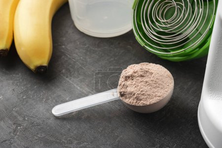 Proteína de suero en polvo con sabor a chocolate en una cuchara de plástico sobre un fondo oscuro con plátanos y una coctelera.