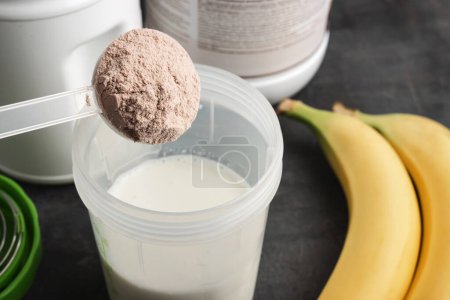 Cuchara de medición de plástico pone proteína de suero en polvo en una coctelera con leche sobre un fondo oscuro con plátanos, proceso de hacer bebida saludable proteína.