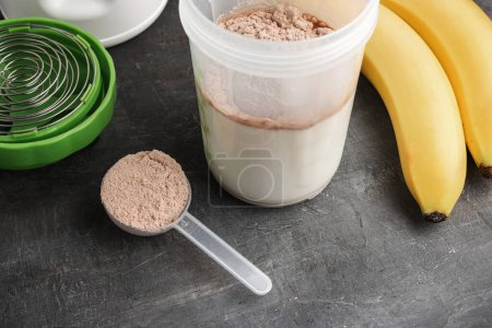 Proteína de suero en polvo con sabor a chocolate en una cuchara de plástico, batidora con leche y plátanos sobre un fondo oscuro, proceso de hacer bebida batida de proteínas.