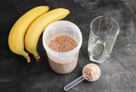 Bebida de proteína de chocolate mezclada en una coctelera, cuchara dosificadora de plástico con polvo de proteína, plátanos y vaso de agua sobre un fondo oscuro.