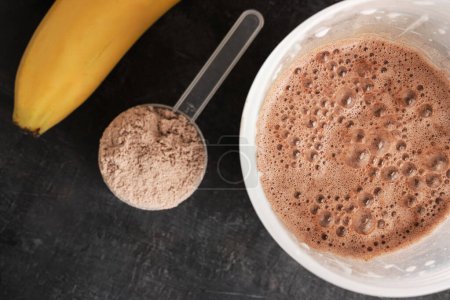 Bebida de proteína de chocolate mezclada en una coctelera, cuchara de plástico con polvo de proteína, plátanos sobre un fondo oscuro, vista superior.