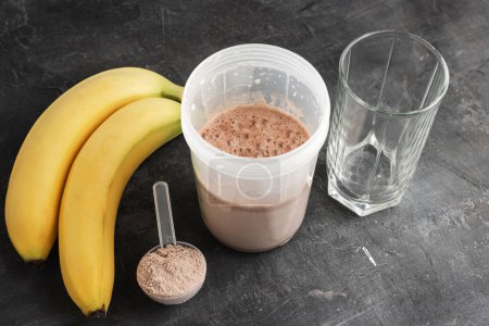 Bebida de proteína de chocolate mezclada en una coctelera, cuchara dosificadora de plástico con polvo de proteína, plátanos y vaso de agua sobre un fondo oscuro.