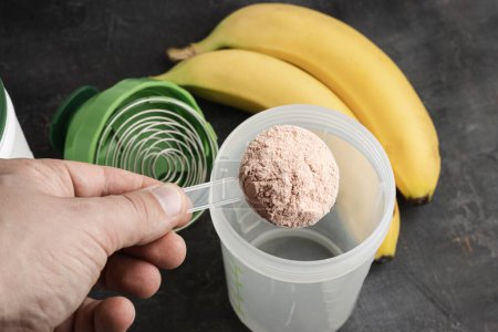 La mano masculina con una cuchara dosificadora de plástico pone la proteína de suero en polvo en una coctelera sobre un fondo oscuro con plátanos, proceso de hacer bebida saludable de proteína.