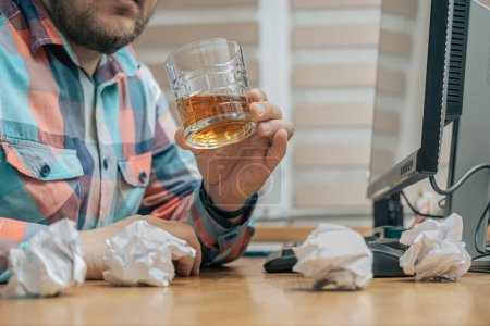 Hombre borracho y frustrado en la oficina mientras trabajaba con PC, sentado en la mesa de trabajo con un vaso de bebida alcohólica, whisky o brandy, alcoholismo y malos hábitos concepto.