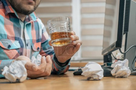 Hombre borracho y frustrado en la oficina mientras trabajaba con PC, sentado en la mesa de trabajo con un vaso de bebida alcohólica, whisky o brandy, alcoholismo y malos hábitos concepto.