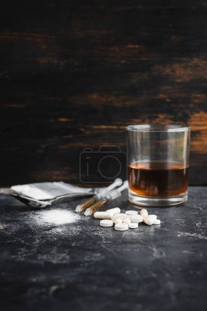 Betäubungsmittel in einem Löffel, Dopingpulver in einer durchsichtigen Plastiktüte, Spritzen mit Medikamentendosis, weiße Pillen und ein Glas Alkoholgetränk auf strukturiertem Hintergrund. Konzept von Sucht und schlechten Gewohnheiten.