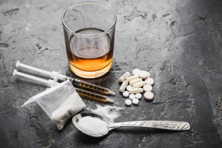Narcotic substances in spoon, dope powder in transparent plastic bag, syringes with drugs dose, white pills and glass of alcohol drink on textured background. Concept of addiction and bad habits.