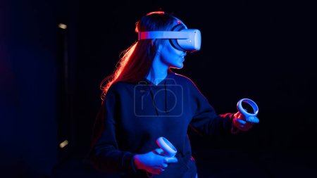 Foto de Una joven mujer emocionada jugando en juegos de realidad virtual usando gafas y controladores de realidad virtual. Iluminación roja y azul - Imagen libre de derechos