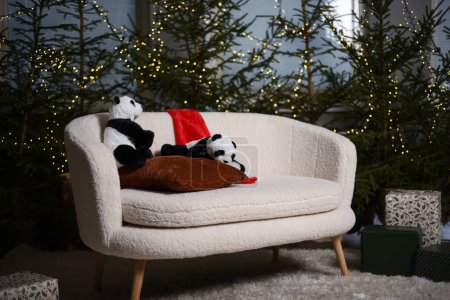 Téléchargez les photos : Intérieur de Noël avec des arbres de Noël verts décorés de lumières derrière un canapé blanc où deux pandas jouet sont assis avec un chapeau d'elfe rouge. - en image libre de droit