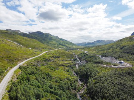 Drone photo. Top view of a Norwegian mountain valley with an asphalt highway going into the distance under a blue sky with cumulus clouds on a sunny summer day.
