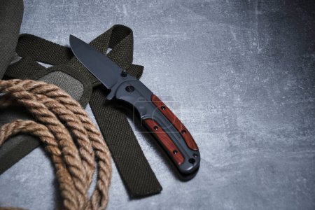 Folding knife for survival, a rope and a khaki backpack strap on a gray concrete background