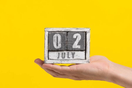 Date is July 02. Wooden calendar in hand on yellow background
