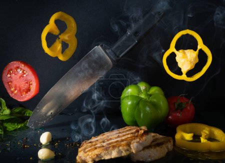 Lévitation alimentaire. Couteau de cuisine, morceaux de légumes lumineux, viande grillée aromatique savoureuse avec fumée chaude sur fond noir Tomate rouge fraîche, vert, poivron jaune, ail, feuilles de basilic, graines de poivre 