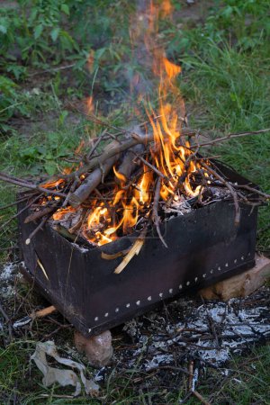 Helles Lagerfeuer vor dem Grillen. Kohlenbecken aus Eisen und trockene brennende Zweige, Holzkohle drinnen auf grünem Grashintergrund. Vertikales Foto.