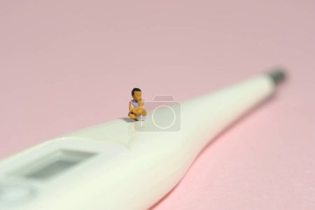 Foto de Miniatura gente pequeña juguete figura fotografía. Una niña pequeño bebé sentado por encima del termómetro mientras gimotea sobre fondo rosa. Foto de imagen - Imagen libre de derechos