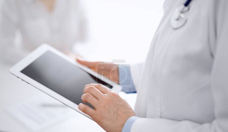 Doctor and patient in clinic. The focus is on female physicians hands using tablet computer at the background of unknown woman. Medicine concept.
