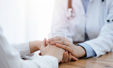 Arzt und Patient sitzen am Holztisch in der Klinik. Ärztinnen reichen der Frau beruhigende Hände. Medizinkonzept.