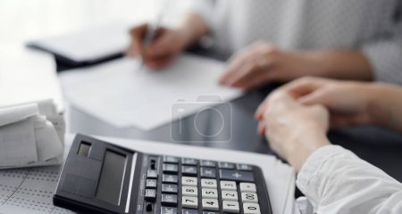 Woman accountant using a pen and laptop computer while counting and discussing taxes with a client, focus on the calculator. Business audit and finance concepts.