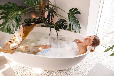 Foto de Mujer joven relajada bañándose con cosméticos para el cuidado del cuerpo en un estante de madera sobre una moderna bañera llena de burbujas blancas en casa. Spa, concepto de bienestar - Imagen libre de derechos