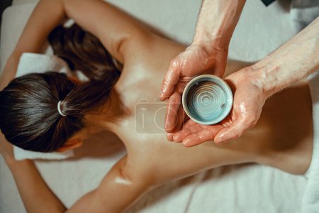 Photo for Masseur man holding bowl with warm herb infused oil for back massage. Woman receiving massage at spa - Royalty Free Image