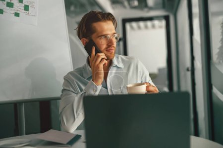 Foto de Hombre jefe hablando de teléfono y beber café mientras está sentado en la oficina moderna con el ordenador portátil - Imagen libre de derechos