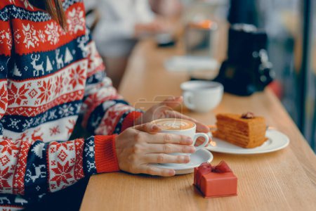 Photo for Young woman enjoying a warm cup of tea in a festive sweater at a charming cafe - Royalty Free Image