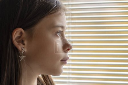 Photo for An 11-year-old girl looks through the lowered blinds of the window at the street, close-up, selective focus. Concept: waiting for parents, orphanage, child support. - Royalty Free Image