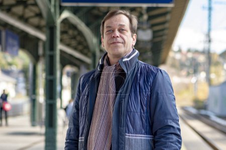 Foto de Retrato de un hombre de 45-50 años de edad en una chaqueta de pie en el andén de la estación de tren, esperando un tren o tren. - Imagen libre de derechos