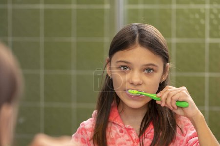 Una chica se cepilla los dientes con un cepillo de dientes en el baño, un reflejo en el espejo, una mirada a la cámara, un plan sobre su hombro.