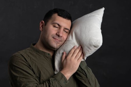 Téléchargez les photos : An emotional portrait of a 30-35-year-old man with his eyes closed, pressing a pillow to his face, sleeping standing on a black background. - en image libre de droit