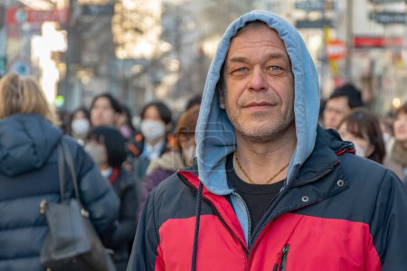 Foto de Street Portrait of a 45-50-year-old man in a hood against the background of a city street and a crowd of people, looking into the camera. - Imagen libre de derechos