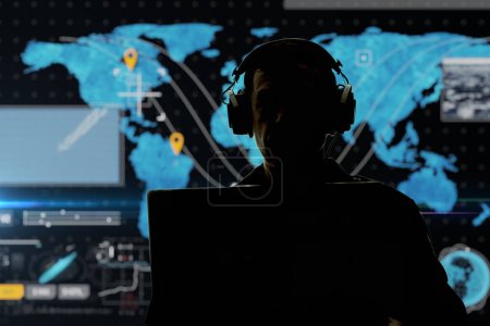 Foto de Silhouette of a military man in headphones at a laptop against the background of a world map, contour lighting. Concept: collection of confidential information, surveillance and control of people. - Imagen libre de derechos