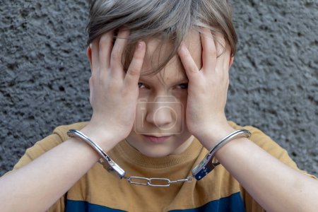 A juvenile delinquent in handcuffs against a gray wall, close-up. Concept: juvenile delinquency, petty theft and theft, incarceration.