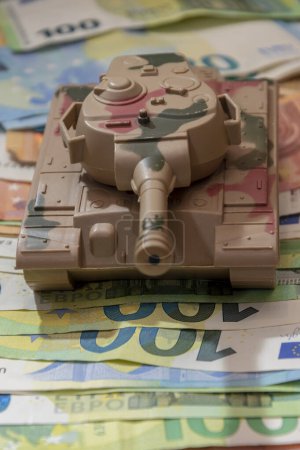 A toy military tank, a scattering of Euro banknotes, a vertical photo. Concept: state budget expenditures on the army and weapons, military assistance to Ukraine, transfer of equipment and weapons.