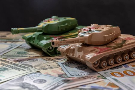 Tanques militares de juguete, dólares americanos dispersos, fondo negro. Concepto: gasto en armas, ayuda militar, comercio de armas y municiones, préstamo de dinero, guerra en Ucrania.