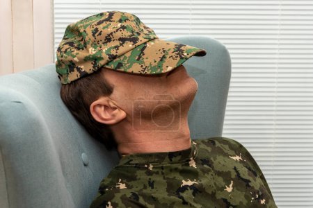 Téléchargez les photos : Un militaire âgé de 45 à 50 ans s'est couvert le visage avec une casquette de l'armée, dormant assis dans un fauteuil. Concept : troubles mentaux chez le personnel militaire, traitement par un psychothérapeute. - en image libre de droit