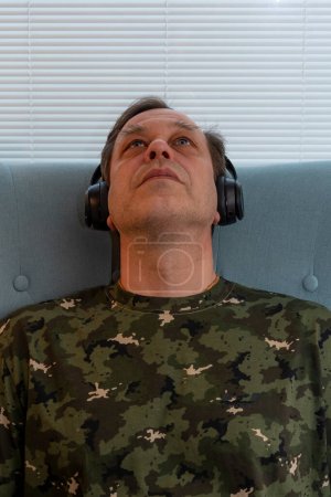 Foto de Retrato de un hombre de uniforme militar de 45-50 años, con auriculares en el fondo de persianas cerradas, escuchando música, psicoterapia para militares. - Imagen libre de derechos