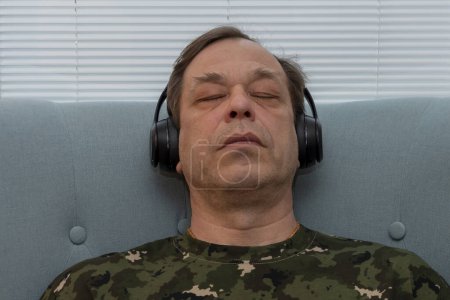 Foto de Un hombre de uniforme militar de 45-50 años con los ojos cerrados, con auriculares en el fondo de persianas, escuchando música, psicoterapia para el personal militar. - Imagen libre de derechos