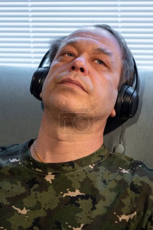 Foto de Un hombre de uniforme militar de 45-50 años, con auriculares en el fondo de persianas de ventana, escuchando música, tratamiento del síndrome postraumático después de la guerra. - Imagen libre de derechos